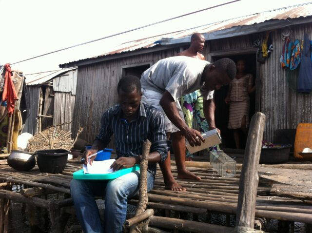 SCARIA researcher taking notes sitting in the frontdoor of a Ladji stilt house while his team-mate set the traps. A lady is looking at them from the house entrance